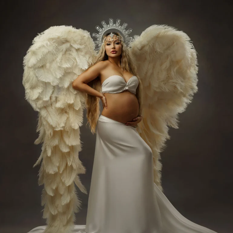 Maternity Photoshoot in North West London 1 Expectant mother in white bandeau and skirt, feathered wings, crown; moody studio portrait.
