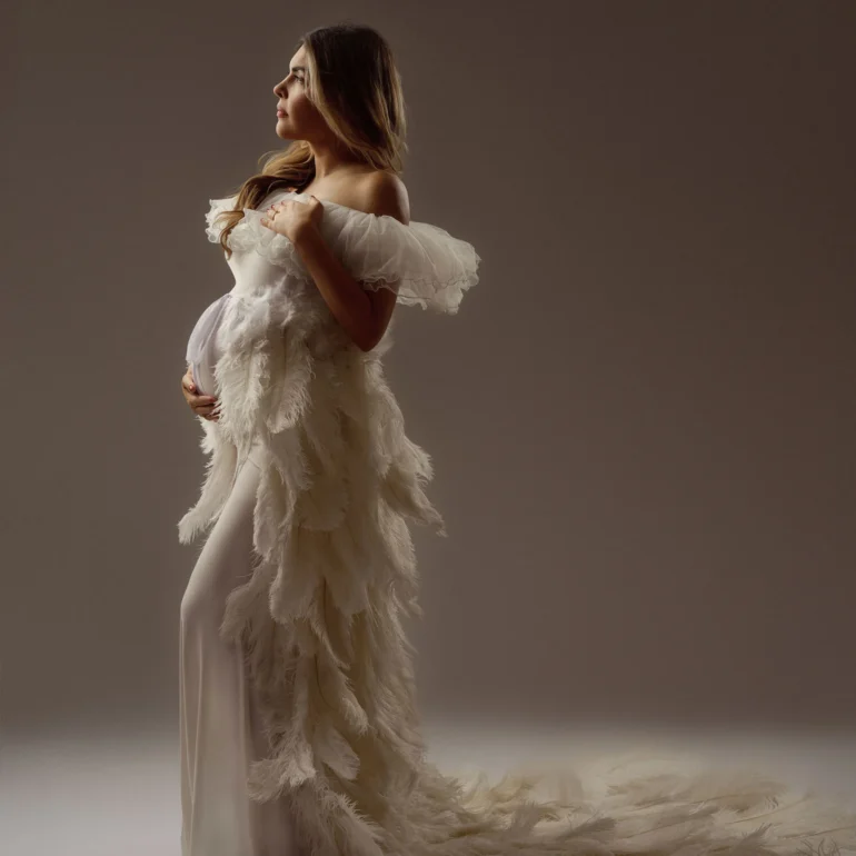 Maternity Photoshoot in North West London 10 Profile in off‑shoulder white feathered gown; soft, ethereal studio scene.