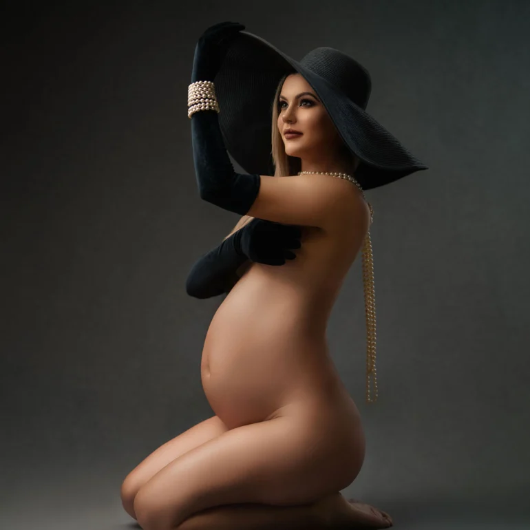 Maternity Photoshoot in North West London 5 Kneeling expectant mother in wide black hat, velvet gloves and pearls; moody backdrop.
