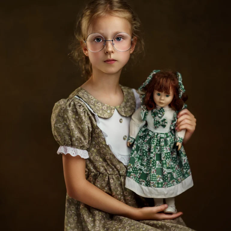 Family & Portrait Photoshoot in North West London 20 Young girl in floral dress holding doll; dark studio backdrop, vintage style.
