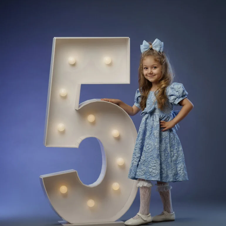 Family & Portrait Photoshoot in North West London 48 Girl in blue dress with bow beside lit number 5; blue studio background.