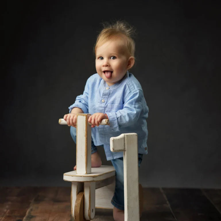 Family & Portrait Photoshoot in North West London 49 Baby boy in light blue on vintage ride‑on with big number 1; playful tongue out.