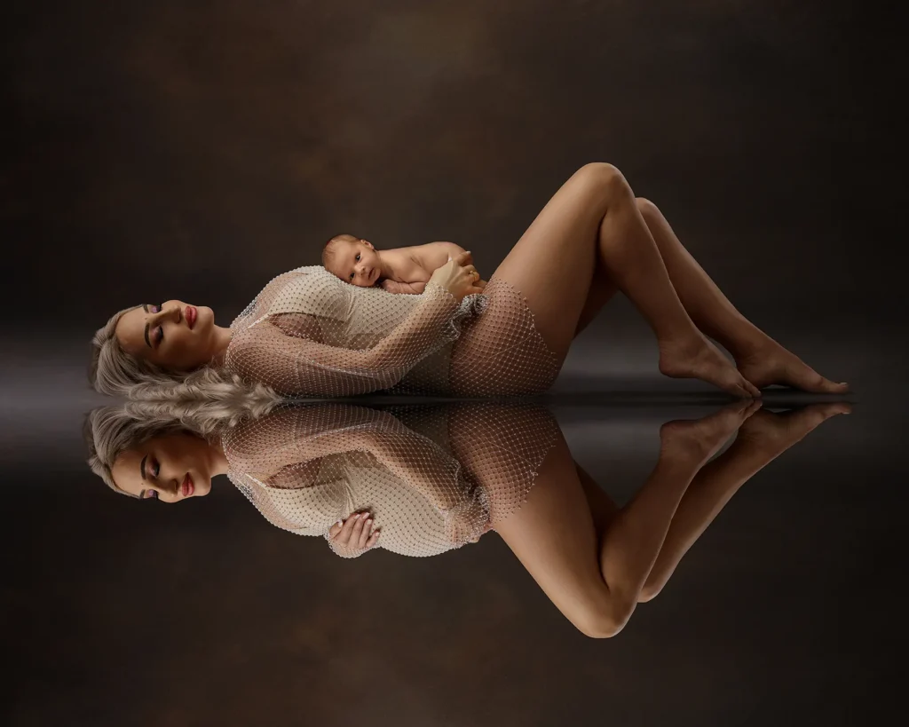 A mother reclines gracefully while holding her newborn baby, captured in a beautifully artistic and reflective composition. The dark, moody backdrop enhances the warmth and connection between them, creating a sophisticated and intimate maternity and newborn portrait.