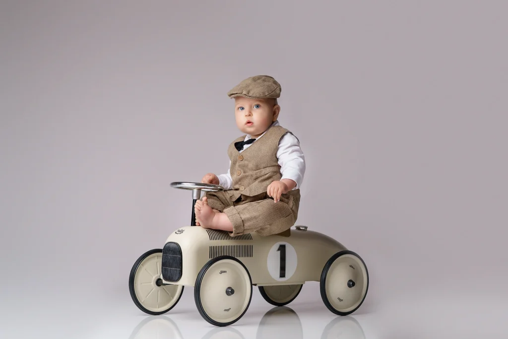 Baby boy in a vintage outfit sitting on a retro-style ride-on car with the number one, captured during a first birthday studio session on a neutral background.